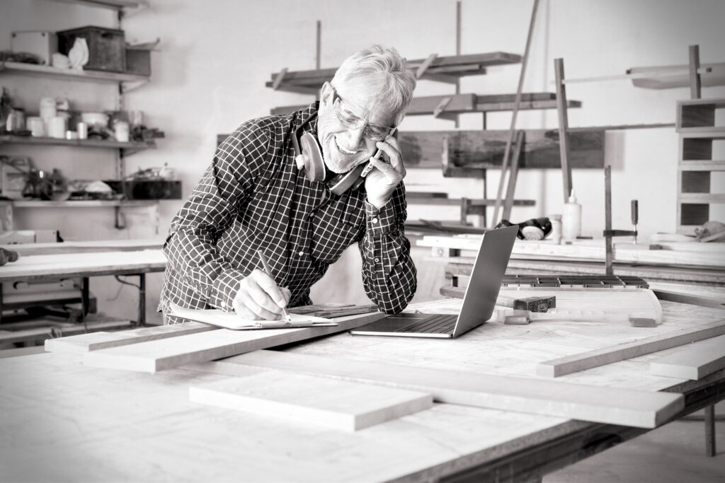 A happy tradesperson smiles while talking into his mobile phone. His other hand is holding a pencil, which he's using to take notes on a clipboard. An open laptop sits on the bench in front of him.