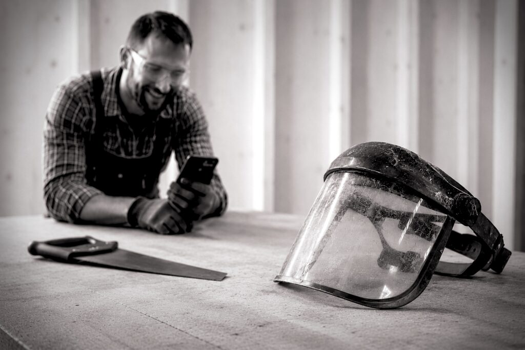 A safety mask / helmet and a hand saw sit on a table. At the far end of the table, a tradesman wearing safety glasses smiles at his phone.