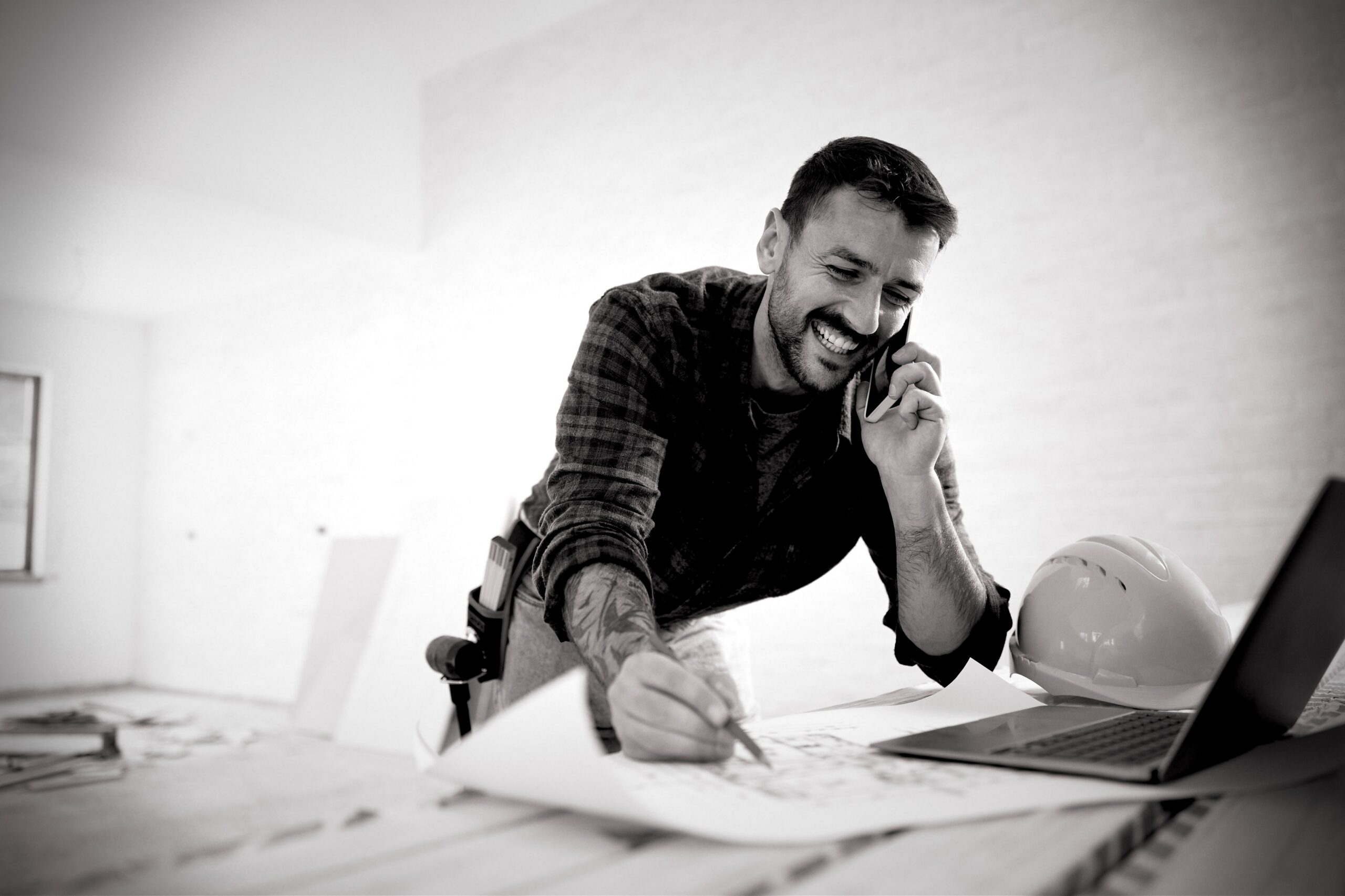 A happy tradesman leans on a workbench while talking into his mobile phone. He's holding a pencil in his other hand, drawing plans for his work. An open laptop and hard hat sit on top of the plans.